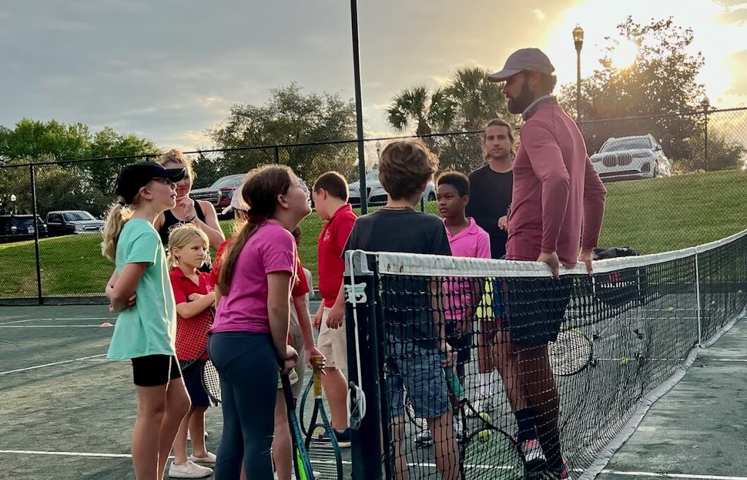 Tennis coach speaks to a group of children on an outdoor court during practice at sunset, nets and rackets visible around them.